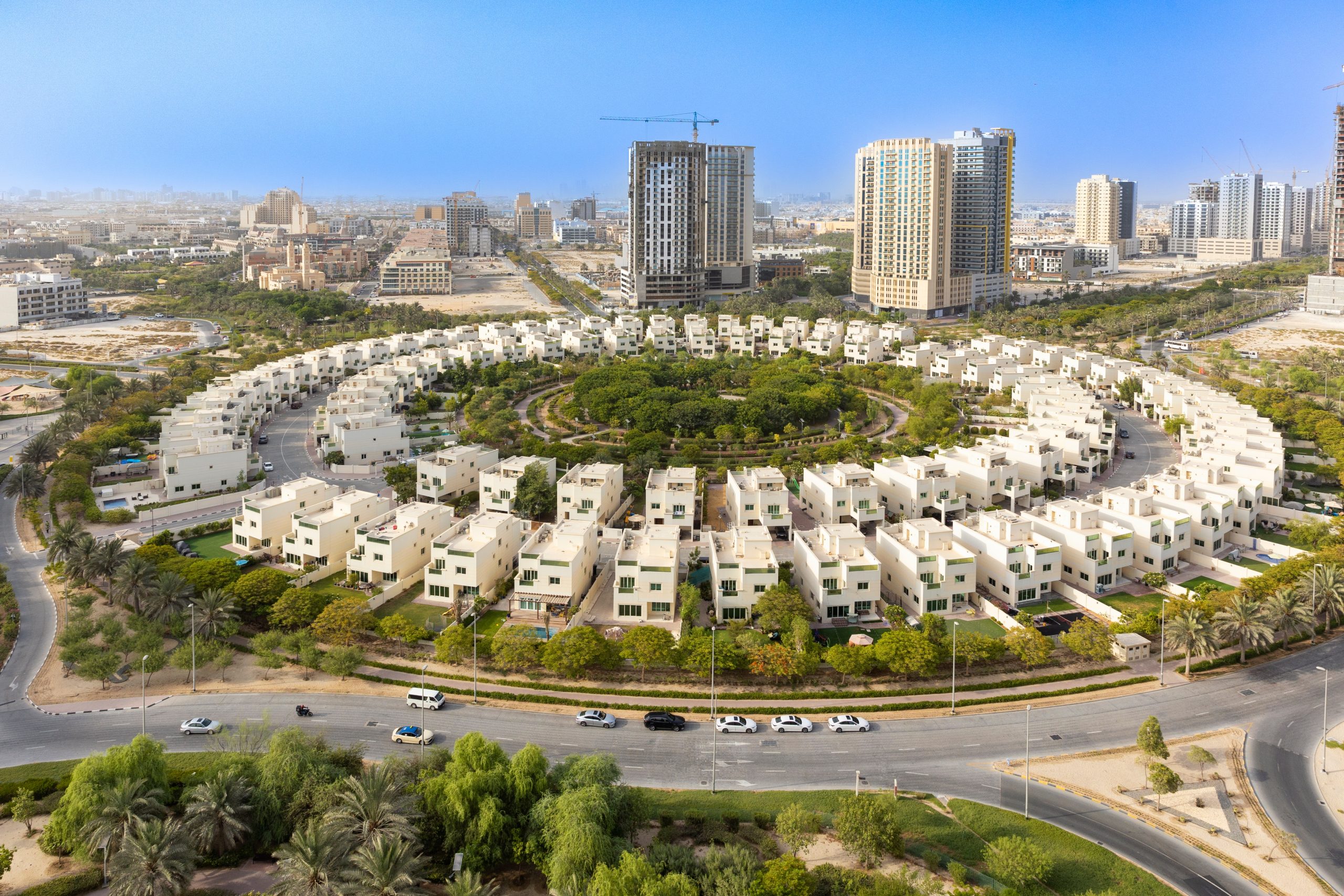 Aerial view of a circular residential community with white houses surrounded by greenery, set amid a modern cityscape with tall buildings and construction cranes in the background.