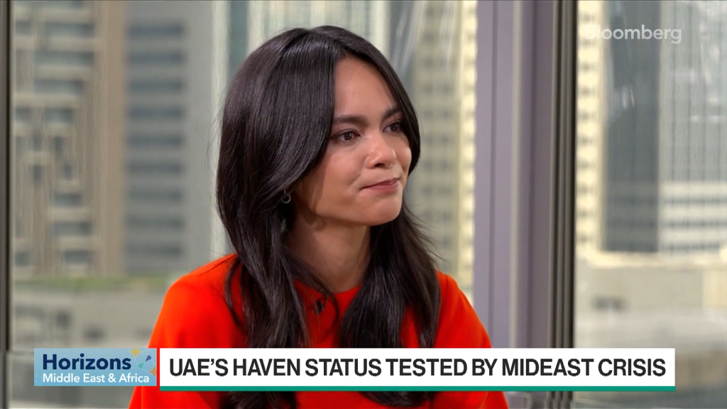 A woman with long dark hair, wearing an orange top, sits indoors against a window with skyscrapers visible. The screen displays Bloomberg graphics and the headline: UAES HAVEN STATUS TESTED BY MIDEAST CRISIS.